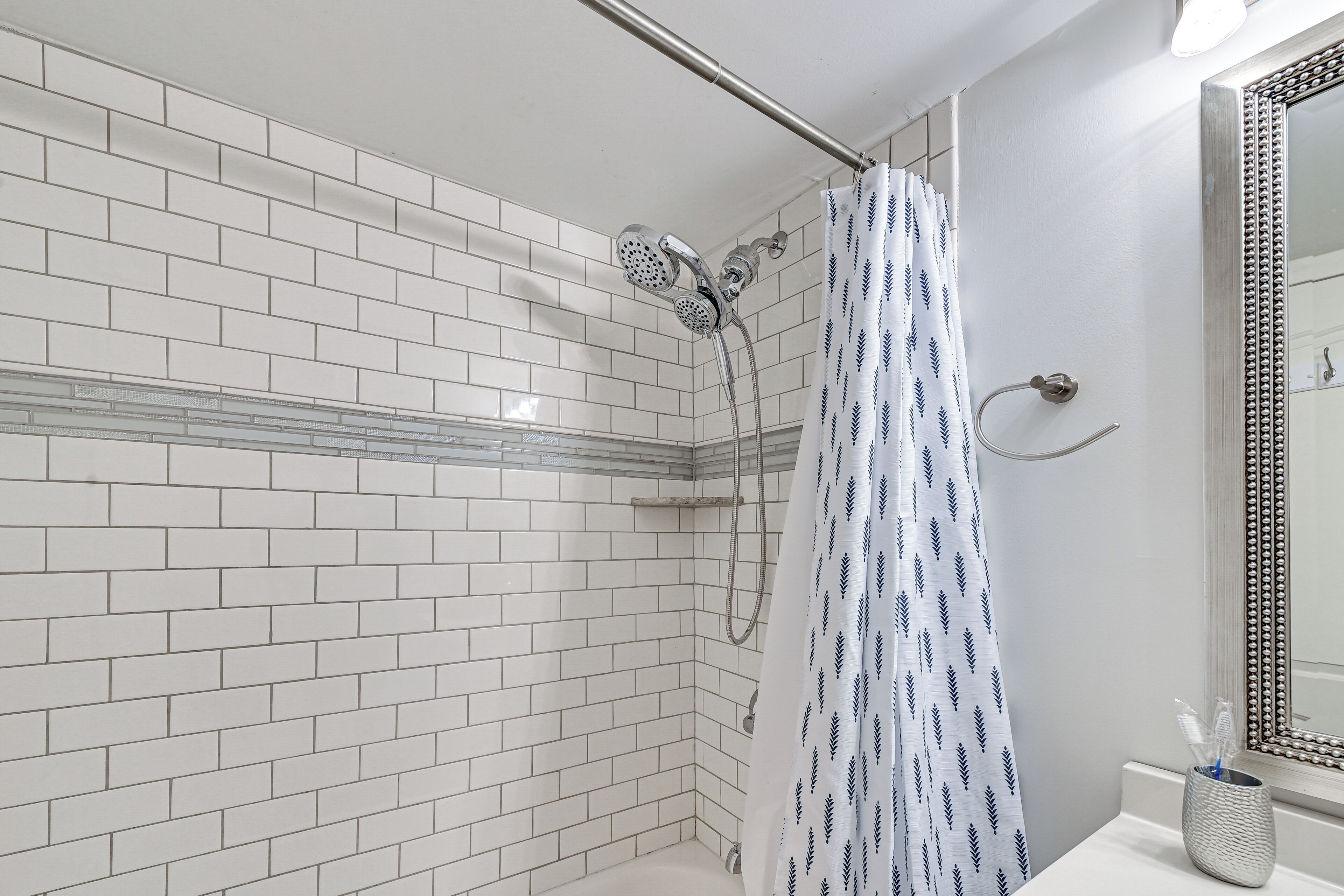 A bathroom with white tiled walls, a shower with a patterned curtain, and a mirror with a decorative frame.