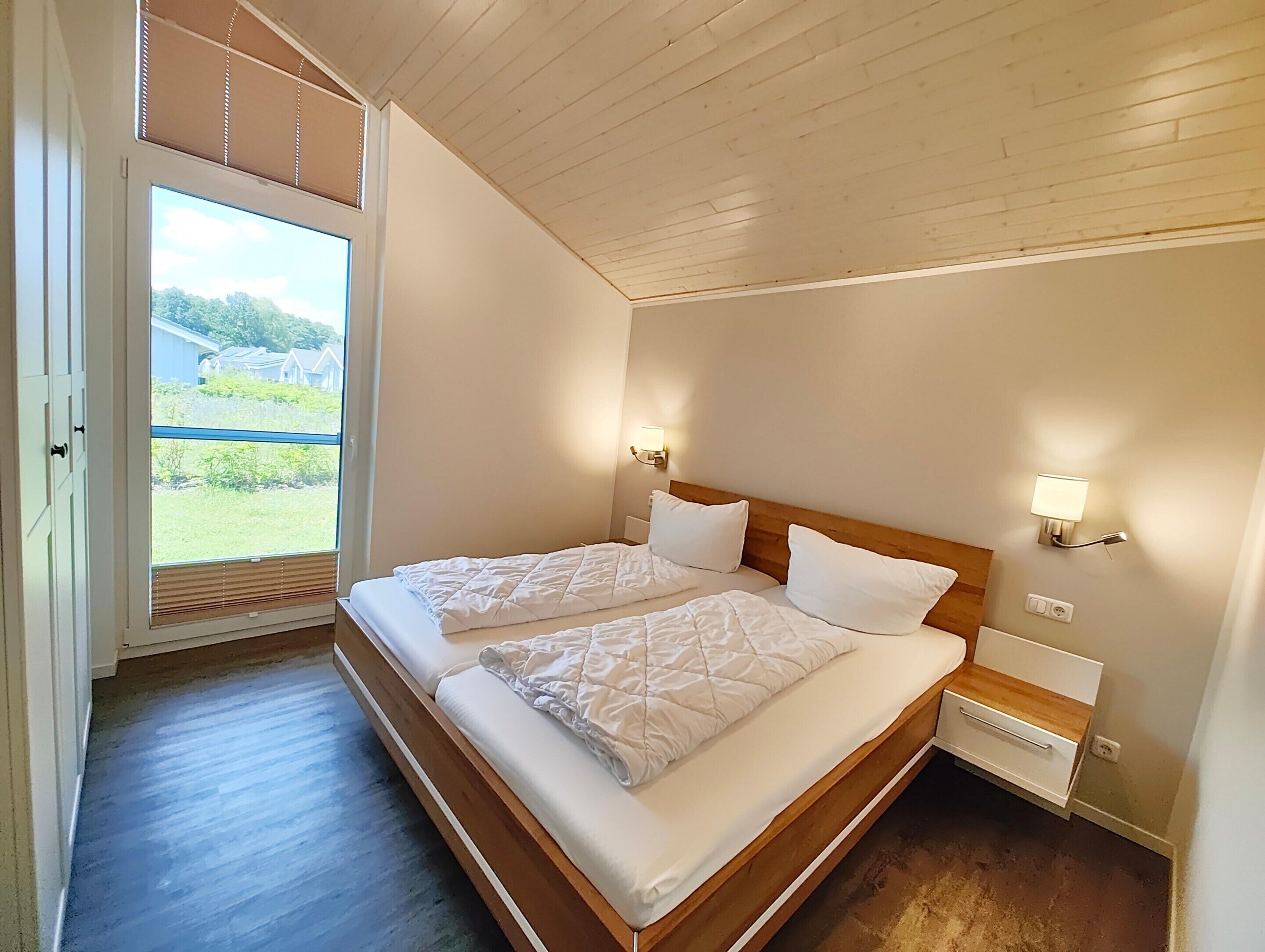A bedroom with two beds, wooden headboards, bedside lamps, and a large window with blinds.