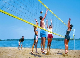 A beach volleyball game with players in swimwear, a net, and a sandy beach with water in the background.