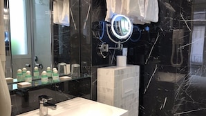Une salle de bain moderne avec un lavabo blanc, une étagère en verre où sont rangés les articles de toilette, un grand miroir doté d'un anneau lumineux et des murs en marbre noir.