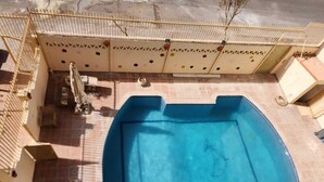 A swimming pool with a tiled deck, a metal fence, and a building with patterned walls in the background. - Villa Yanny Duplexes Apartment Hurghada El Dahar El Helal (Hurghada)