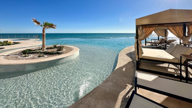 A clear pool with a view of the sea, a palm tree, and a covered lounge area with chairs. - Paradise Found Luxe 2BR Condo in Iconic Location (Puerto Peñasco)