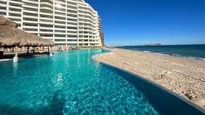 A hotel with a pool extending to the beach, clear blue water, and a thatched-roof structure. - Paradise Found Luxe 2BR Condo in Iconic Location (Puerto Peñasco)