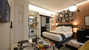 A hotel room with a bed, a dining table set with plates and glasses, a washing machine, and a brick wall with framed pictures.
