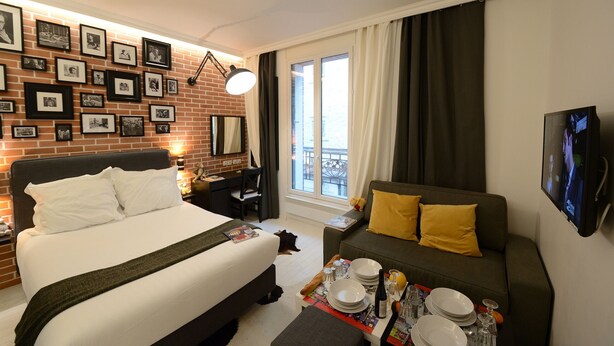 A hotel room with a bed, a sofa, a dining table set with plates and glasses, a TV, and a wall with framed pictures.