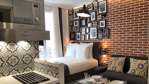 Ein modernes Hotelzimmer mit einer Küchenzeile, einem Bett mit weißer Bettwäsche, einer Ziegelwand mit gerahmten Bildern und einer Sitzecke mit einem gemusterten Sofa und Poufs.