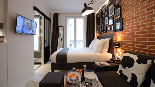 Une chambre d’hôtel moderne avec un grand lit, une table à manger avec des éléments pour le petit-déjeuner, une télévision et un mur en briques orné de photos encadrées.