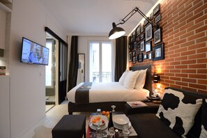 A modern hotel room with a large bed, a dining table with breakfast items, a TV, and a brick wall with framed photos. - Voûte 303 (París)
