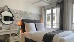 Une chambre avec un lit, un bureau, une chaise, un grand miroir et un papier peint représentant un paysage urbain.