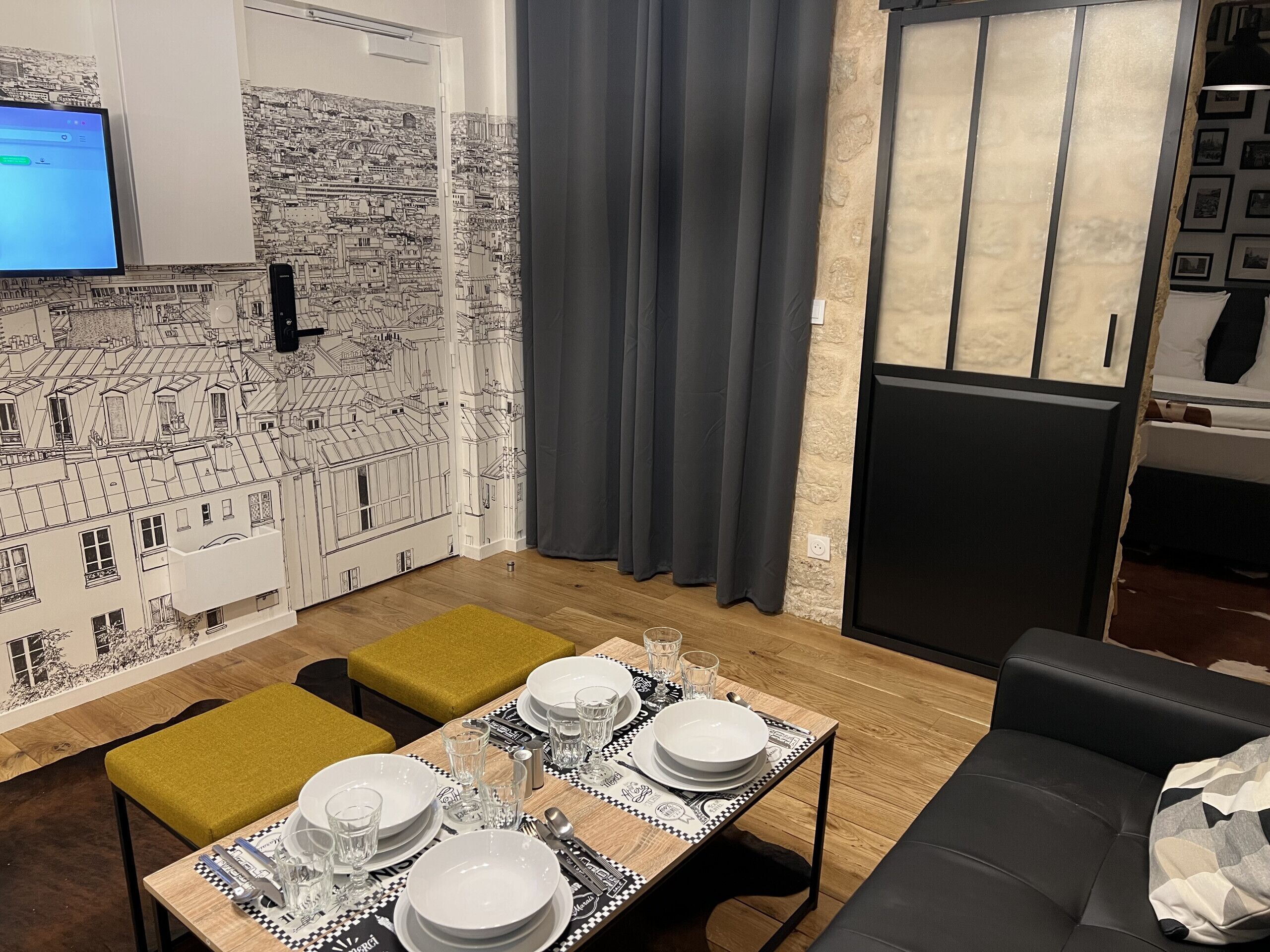 A dining area with a table set for four, a cityscape mural on the wall, and a television mounted above the door.
