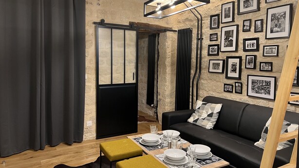 A modern living room with a black sofa, a dining table set with plates and glasses, a wooden coffee table, and a wall adorned with framed photographs. - Loft Jaures (París)