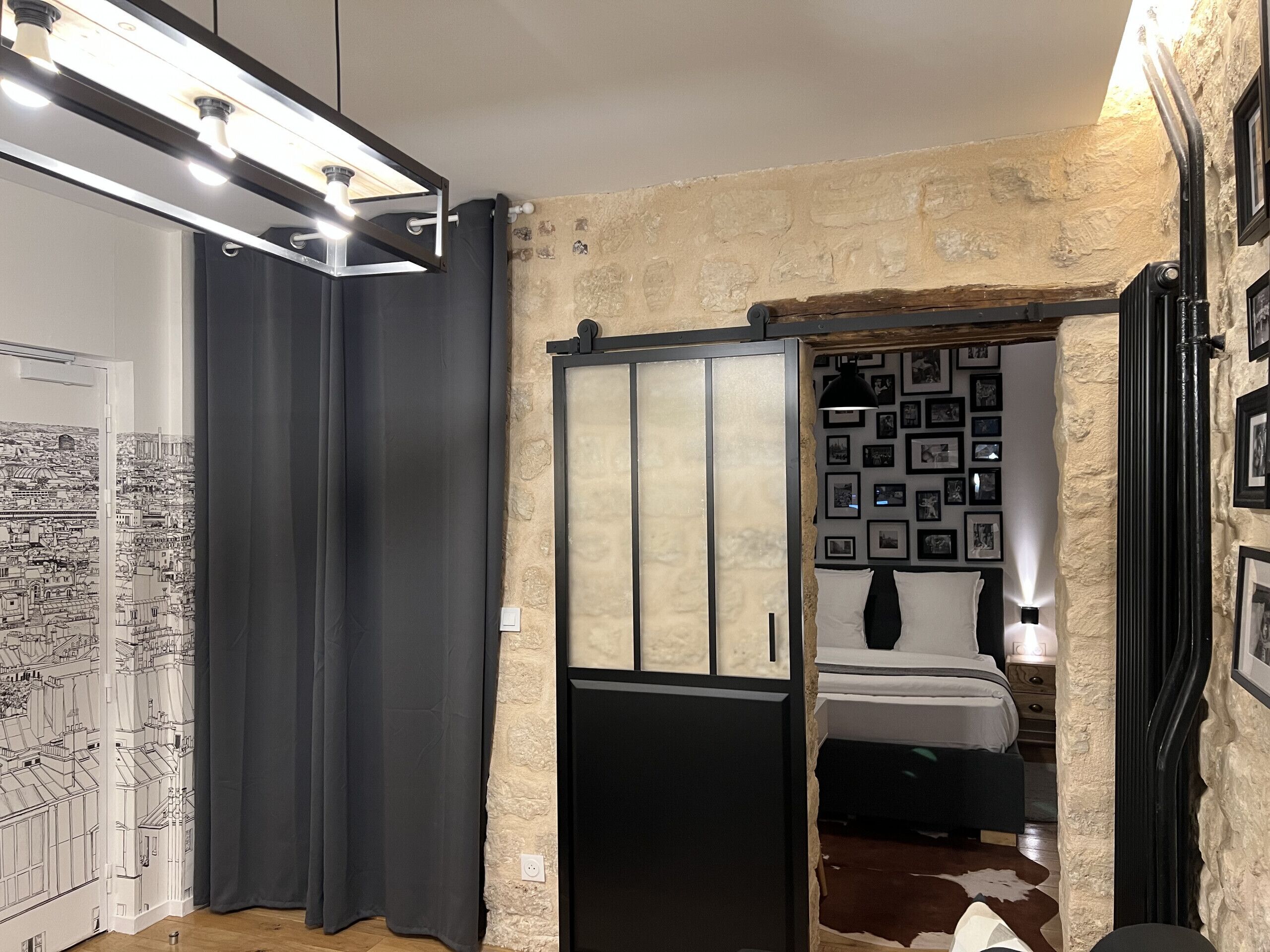 Une pièce avec une grande fenêtre, un lit doté d’une tête de lit blanche, un mur orné de tableaux encadrés et une porte avec un rideau.
