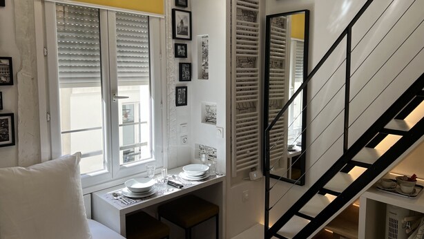 A dining area with a table set for two, a window with blinds, and a staircase with a black railing.