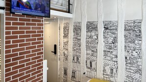A room with a brick wall, a television displaying a news broadcast, and a large black and white cityscape mural on the wall.