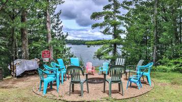 A lakeside seating area with colorful Adirondack chairs arranged around a fire pit, surrounded by trees and a lake view.