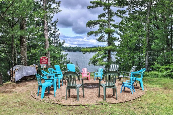 Un espacio de descanso junto al lago con sillas Adirondack de colores dispuestas alrededor de una chimenea, rodeado de árboles y vistas al lago.