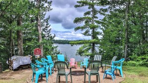 A lakeside seating area with colorful Adirondack chairs arranged around a fire pit, surrounded by trees and a lake view. - The Pointe Resort & Club 1 Bedroom Condo (Minocqua)