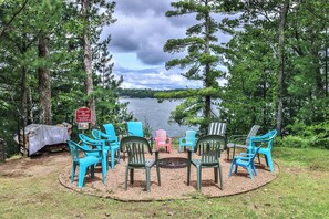 A lakeside seating area with colorful Adirondack chairs arranged around a fire pit, surrounded by trees and a lake view.