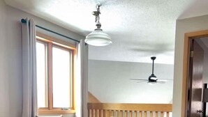 Zona pranzo con tavolo in legno e quattro sedie, una ventola a soffitto e vista su un balcone attraverso una finestra.