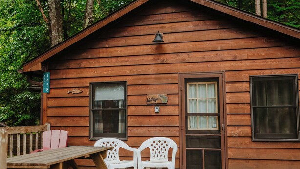 Une cabane en bois avec une table de pique-nique et deux chaises en plastique blanc à l’extérieur.