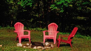 Two red Adirondack chairs arranged around a campfire in a grassy area with trees in the background. - Old Stoney's Walleye- PET FRIENDLY (Arbor Vitae)