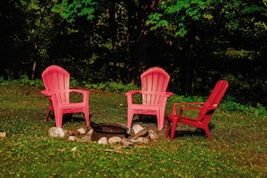 Two red Adirondack chairs around a campfire in a wooded area.