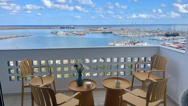 A balcony with four chairs and two round tables, overlooking a marina with boats and a clear sky. - C'est La Vue - Beachfront luxurious condo in Rethymno, Crete (Rethymno)