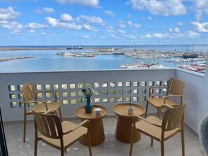 Un balcon avec quatre chaises et deux petites tables rondes, donnant sur une marina où sont amarrés des bateaux, et sous un ciel dégagé.
