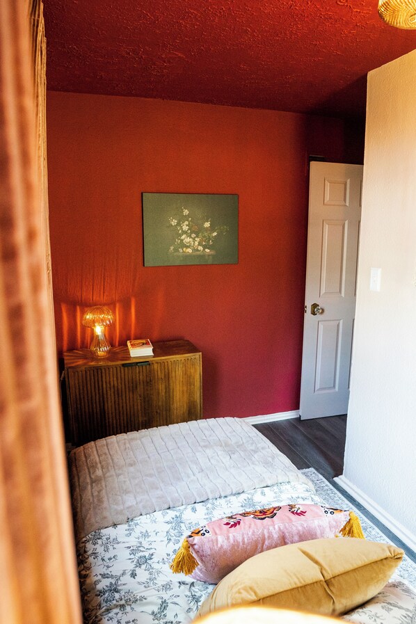 Un dormitorio con cama, mesita de noche con lámpara, puerta y pared con un cuadro enmarcado.