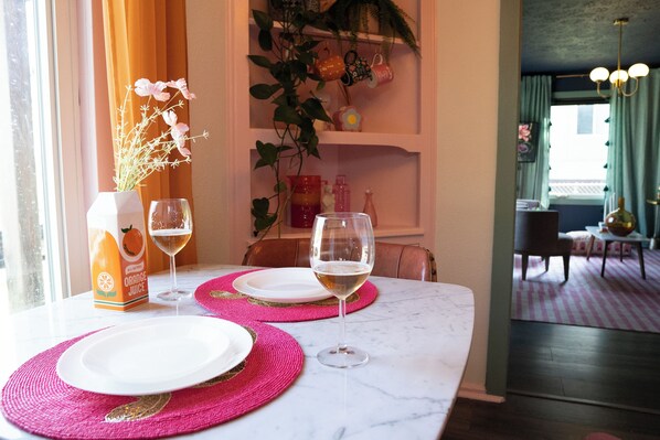 A dining table set with white plates, pink placemats, and two glasses of white wine, with a bottle of orange juice and a vase of flowers on the table.