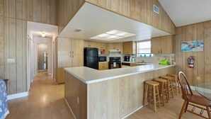 A modern kitchen with wooden paneling, a central island with stools, and a dining area with a glass table. - The Twins: Your Perfect Budget Beach Getaway! (Anna Maria)