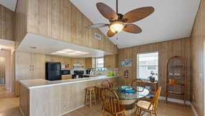 A modern kitchen with wooden cabinetry, a central island, and a dining area with a round table and chairs. - The Twins: Your Perfect Budget Beach Getaway! (Anna Maria)