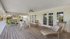 A covered porch with wooden flooring, white walls, and a ceiling fan. There are white chairs and a striped sofa with cushions. - The Twins: Your Perfect Budget Beach Getaway! (Anna Maria)