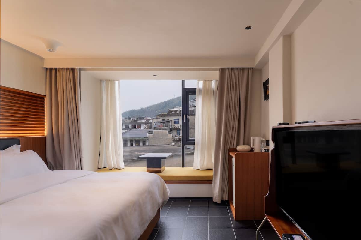 Et hotelværelse med en seng, fjernsyn, et lille bord og et vindue med gardiner.
