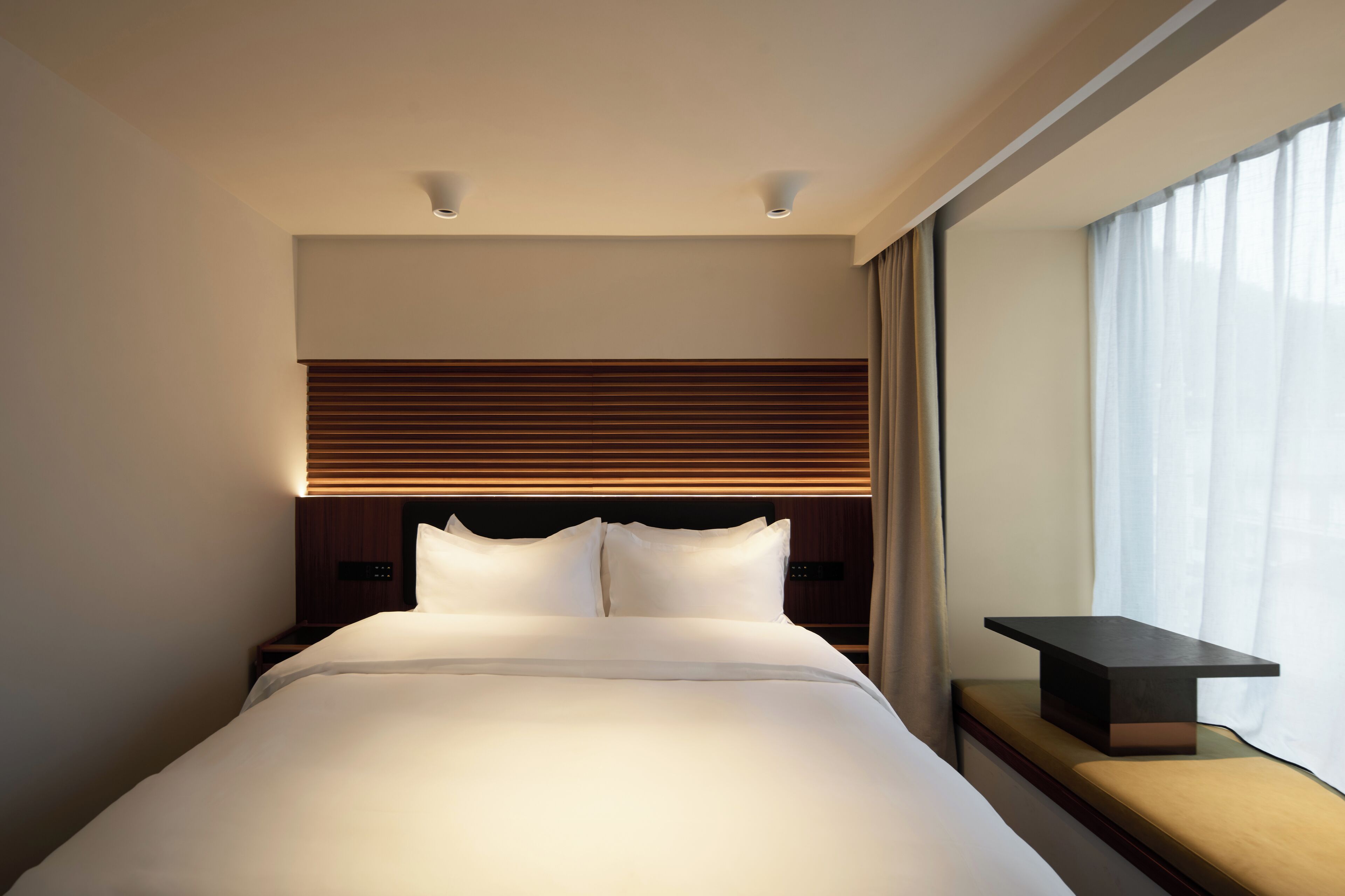 Una habitación de hotel moderna con una cama grande, un cabecero de madera, una mesita de noche y una ventana con cortinas.