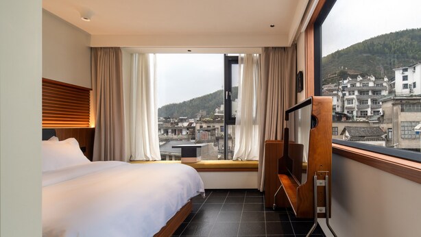 A hotel room with a bed, a window with curtains, a TV, and a view of a cityscape. - The Nest 1219 (Huangshan)