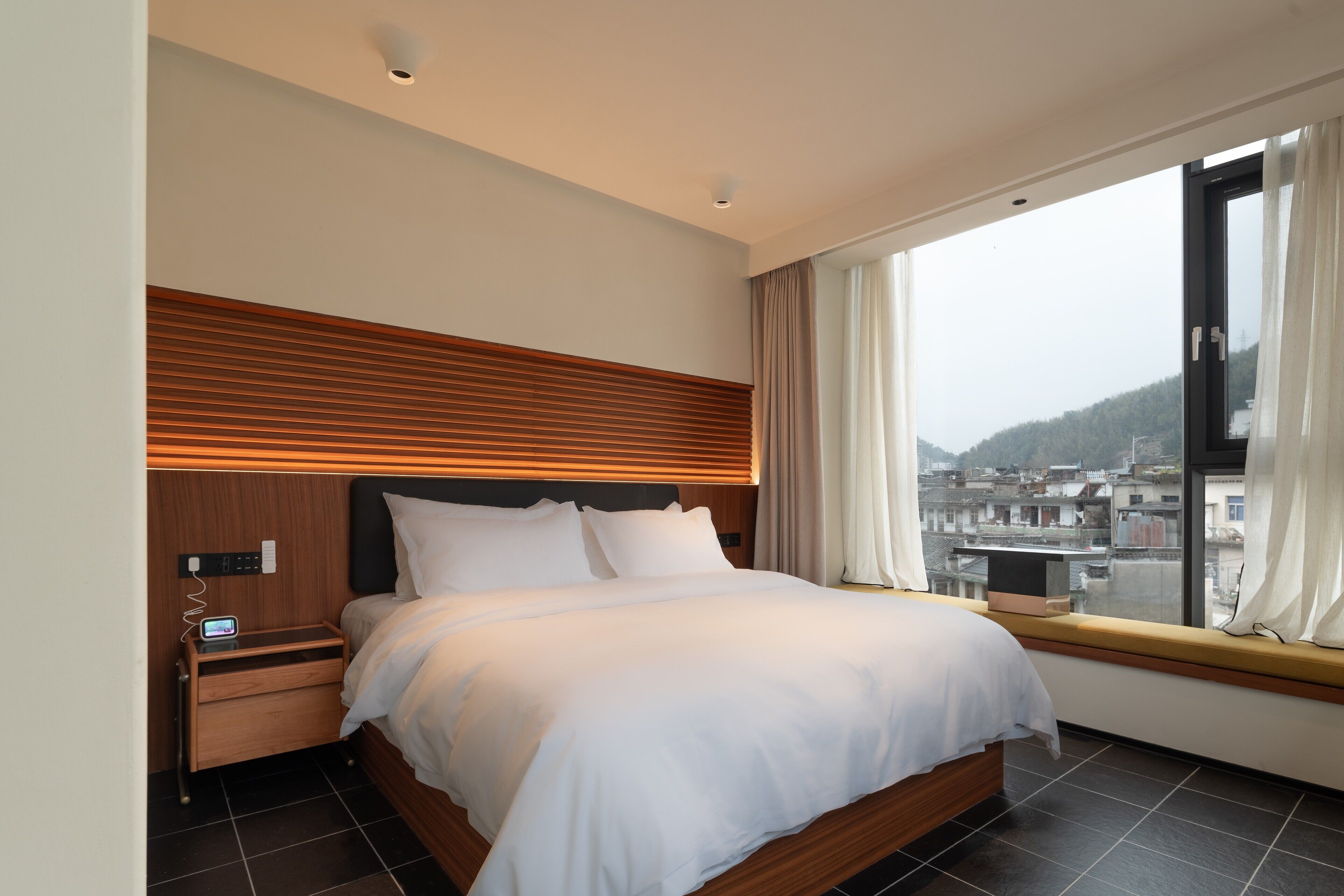 A modern hotel room with a large bed, wooden headboard, and a view of the city through a large window.