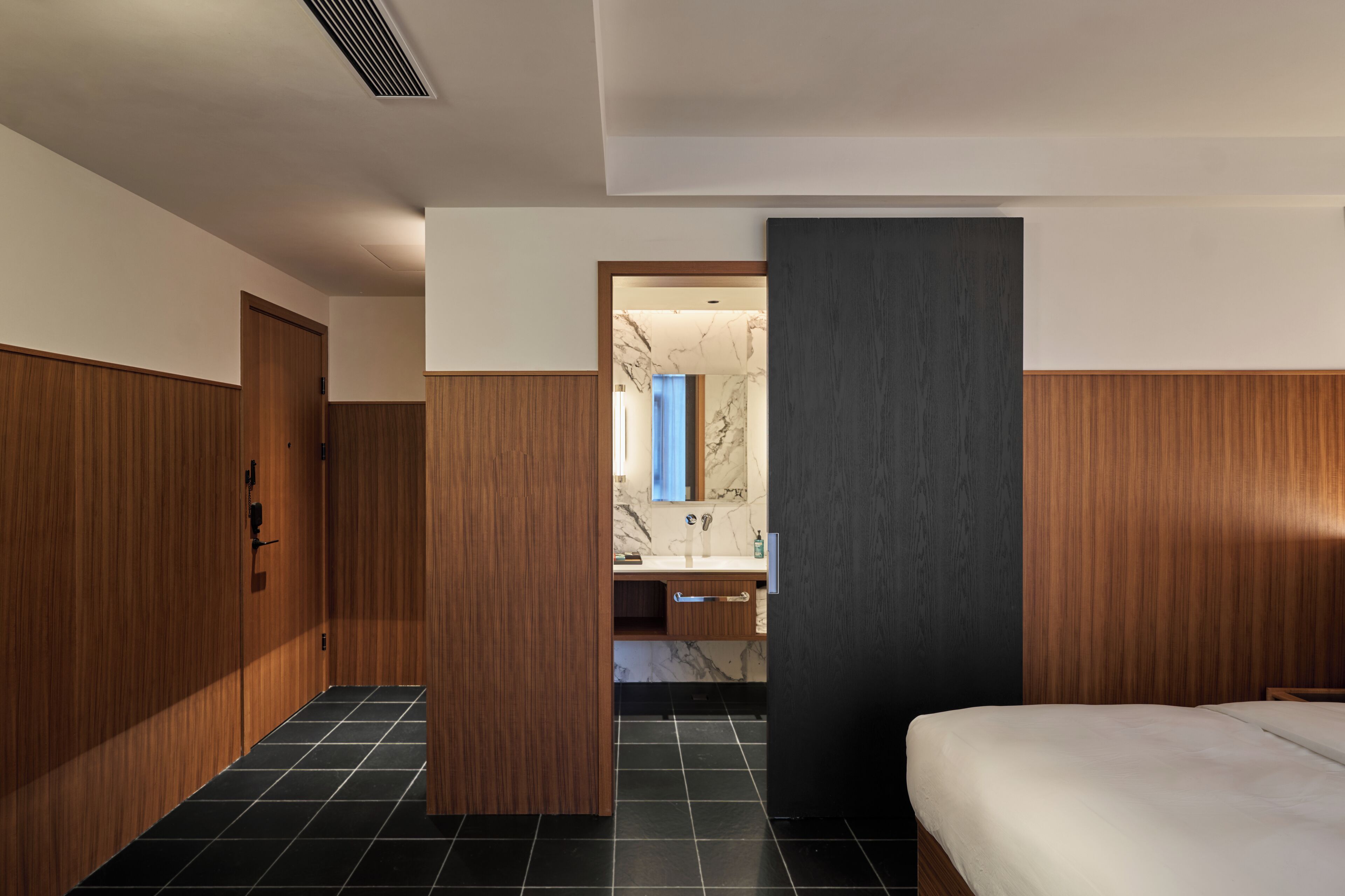 A modern hotel room with a wooden wardrobe, a marble-tiled bathroom, and a bed with a dark headboard.