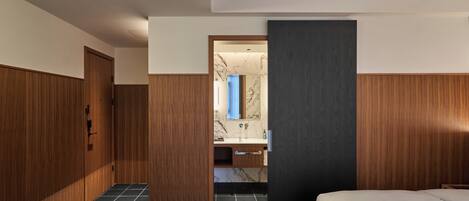 A modern hotel room with a wooden wardrobe, a marble-tiled bathroom, and a bed with a dark headboard.