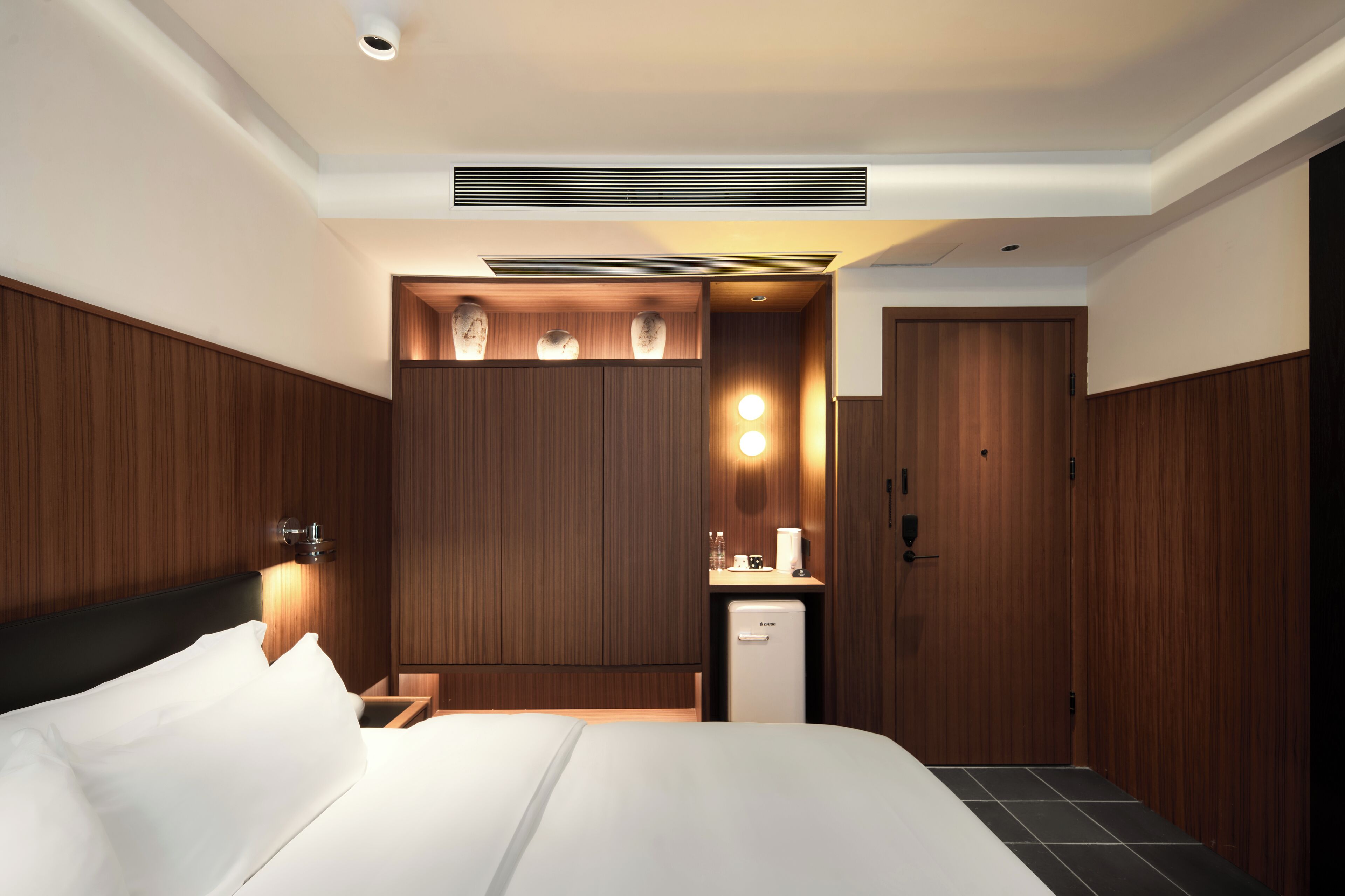 A modern hotel room with a large bed, a small kitchenette, and a wardrobe.