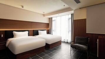 Habitación de hotel con dos camas, una silla y un ventanal grande con cortinas.