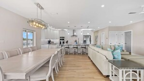 A modern kitchen with white cabinets, a dining area with a long table and chairs, a central island with bar stools, and a light-colored sofa with decorative pillows. - Sunset Point | Luxury 5 bdrm w pool, jacuzzi and XL dock, walk to beach (Marathon)