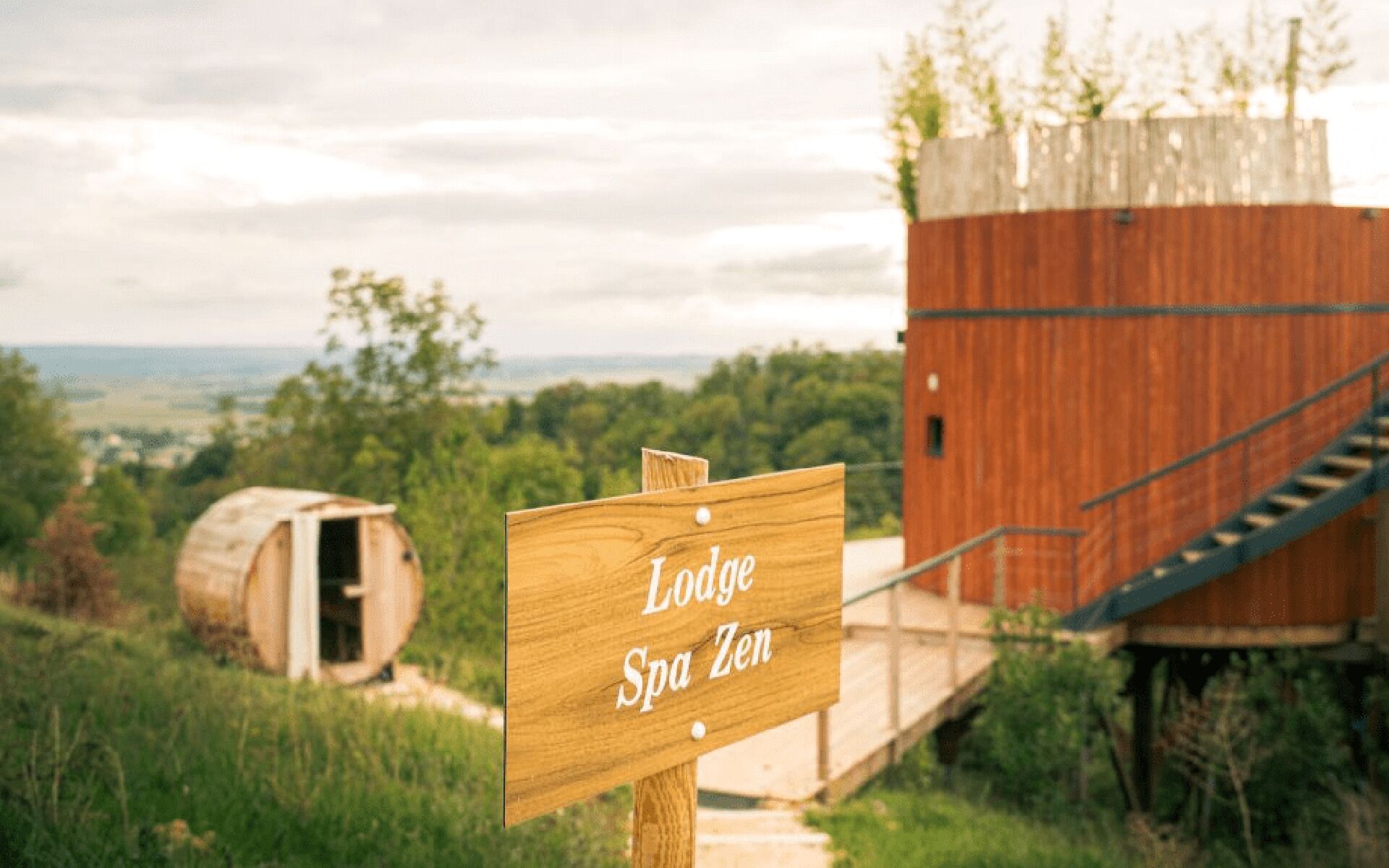 A wooden sign with 'Lodge Spa Zen' written on it, a wooden structure with a staircase, and a barrel in the background.