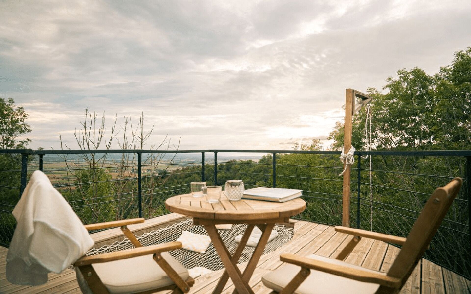 A wooden deck with a round table and two chairs, overlooking a landscape with trees and a fence.