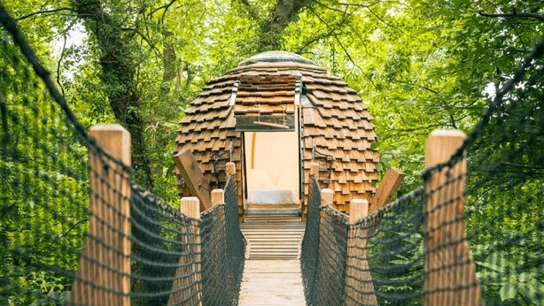 Eine Holzbrücke führt zu einem Bauwerk mit einer kuppelförmigen Dachkonstruktion, umgeben von dichtem Grün.