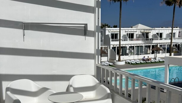 A balcony with white chairs and a small table, overlooking a pool and a resort building. - Tiger’s Eye, a luxurious apartment situated in the heart of Puerto del Carmen (Puerto del Carmen)