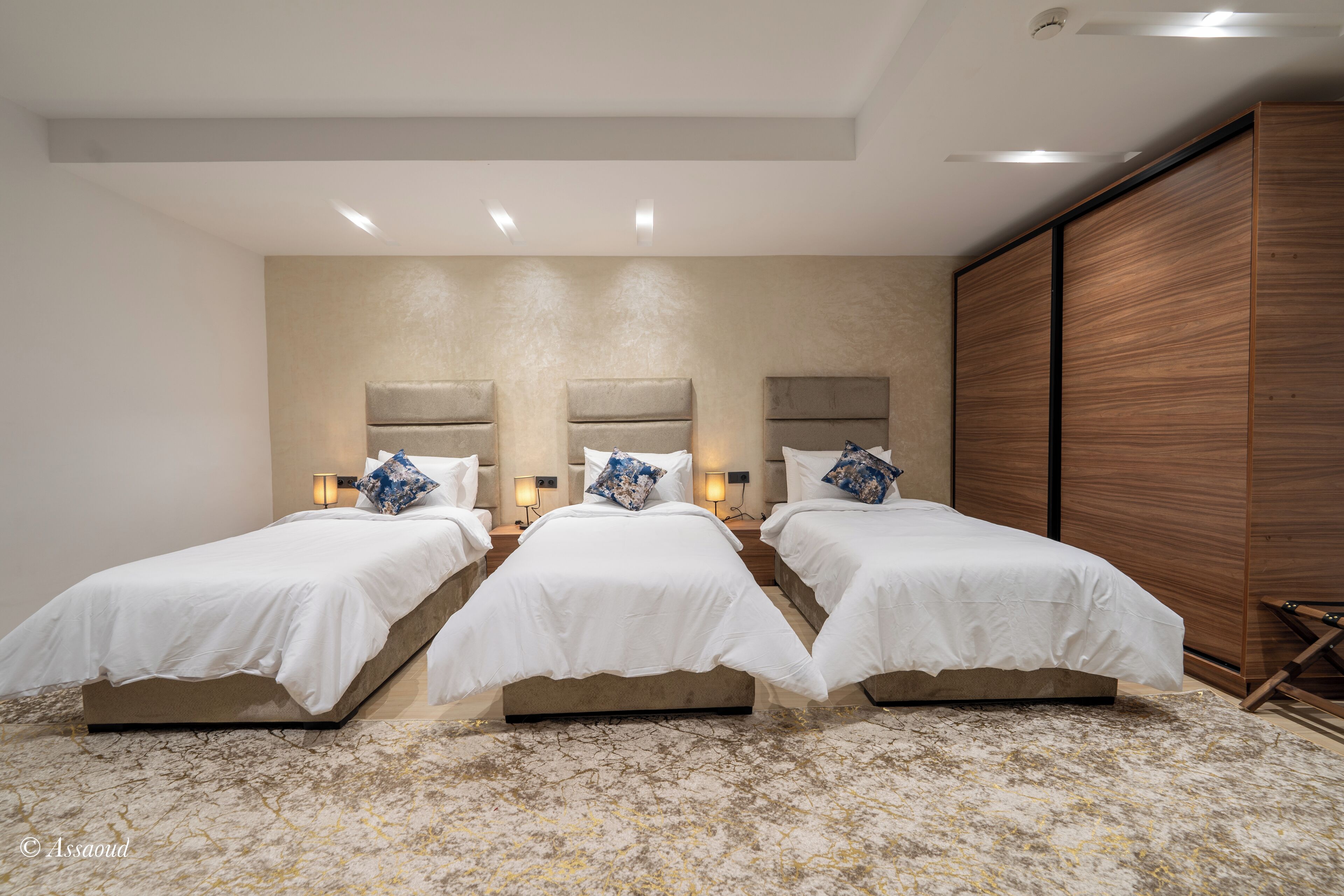 A hotel room with three beds aligned in a row, each with a headboard and bedside lamps.