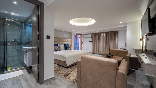 A modern hotel room with a glass-enclosed shower, a large bed with a headboard, a seating area with a sofa and armchairs, and a desk with a flat-screen TV. - LUXURY CHAOUEN (Chefchaouen)
