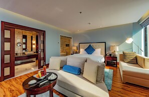 A modern hotel room with a large bed, a sofa, a dining table with fruit, and a view of the city. - Corp Al Khobar Hotel (Al Khobar)