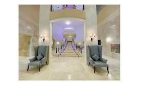 A grand lobby with a central staircase, marble flooring, and two armchairs on either side.